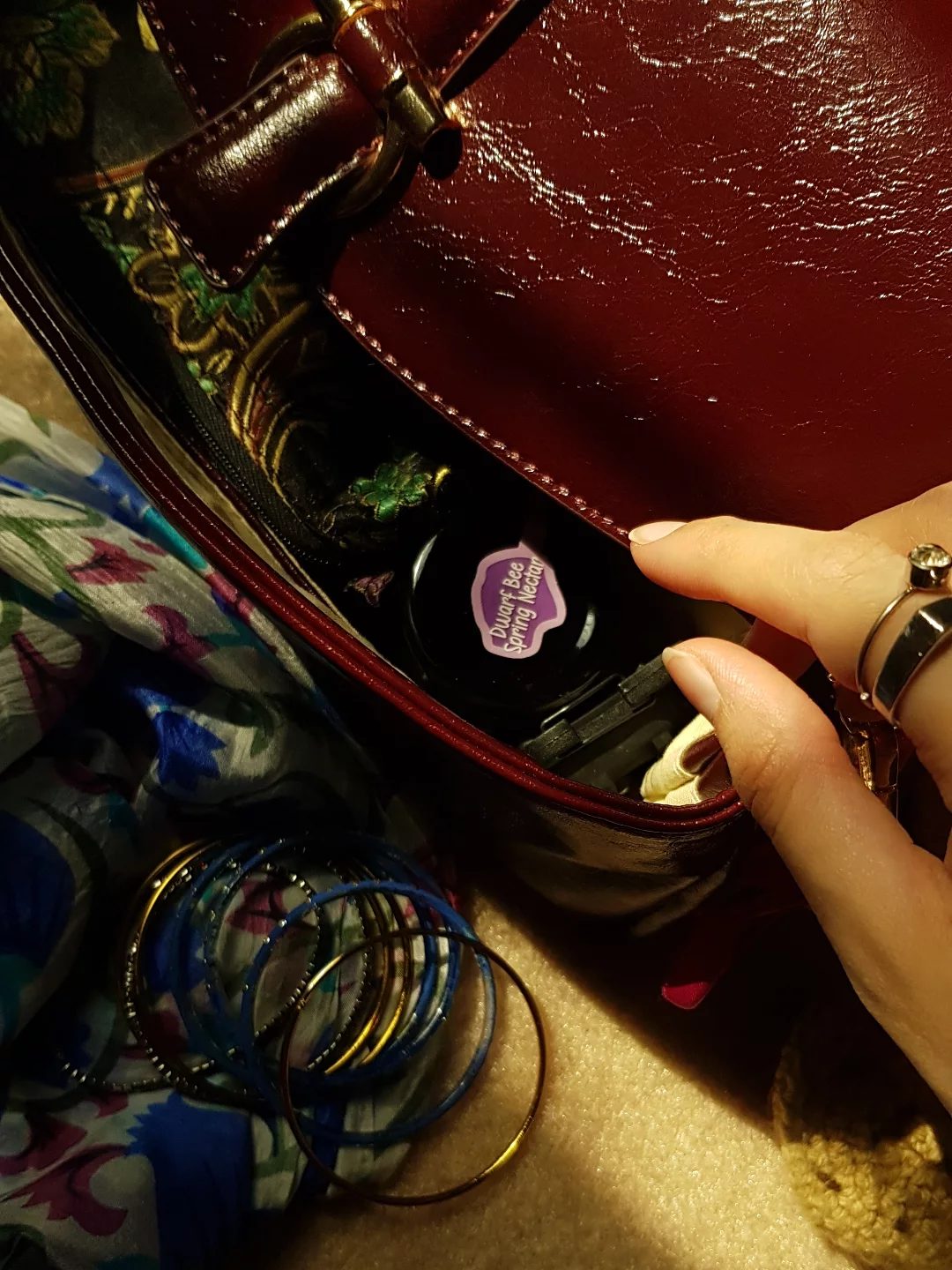 An Asal MINI jar with a purple Dwarf Bee Spring Nectar tab, tucked inside a handbag beside glass bangles and a floral dupatta.